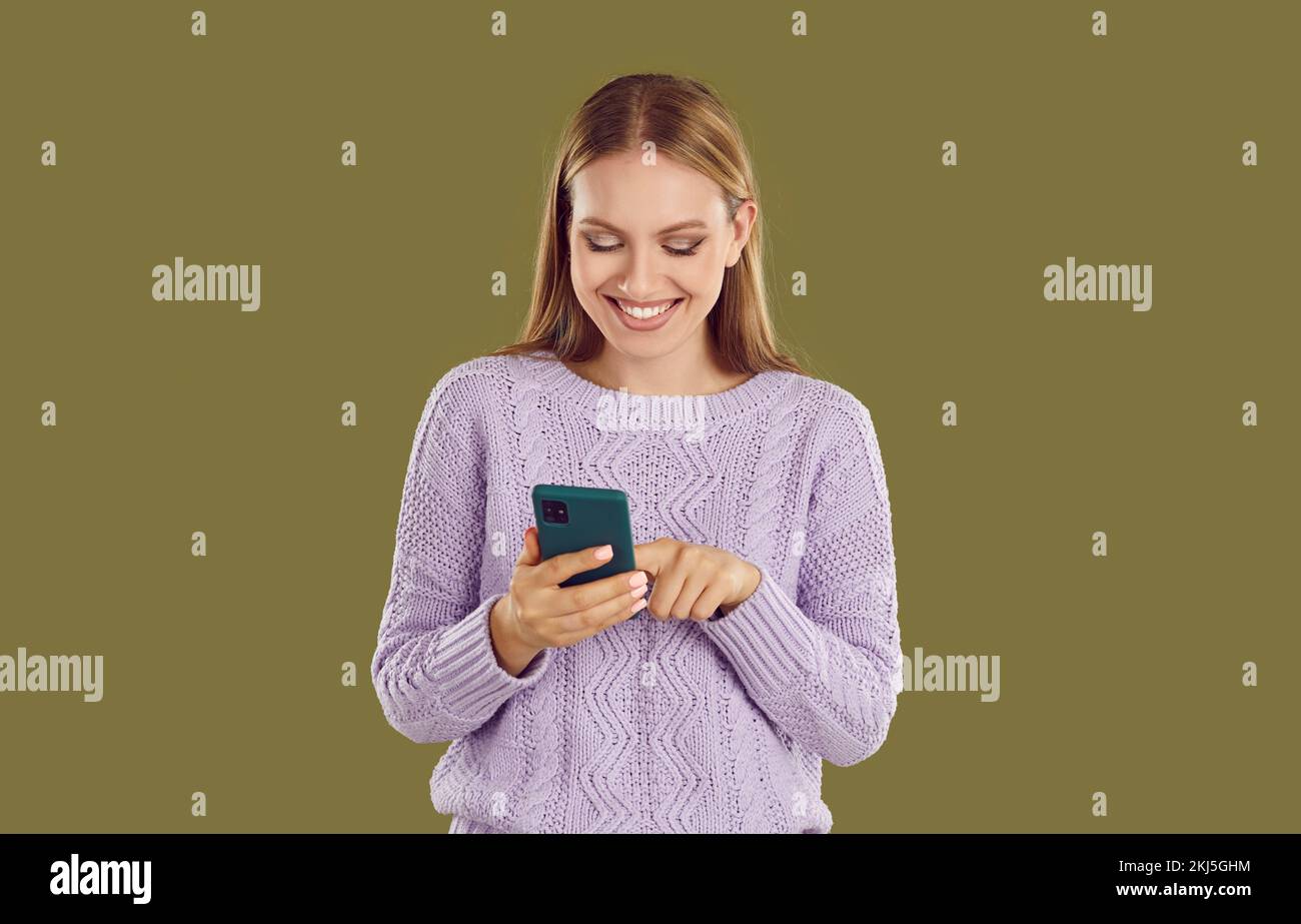 Happy young girl in purple sweater looking at smartphone screen typing message on khaki background. Stock Photo