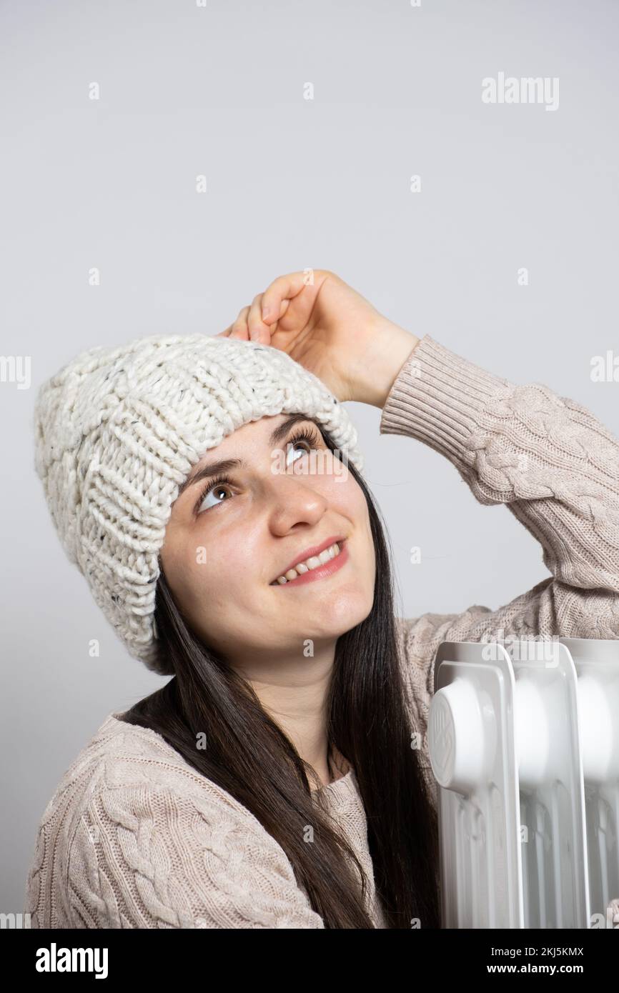 A brunette woman in a hat hugs an oil heater, warms up in winter. Stock Photo