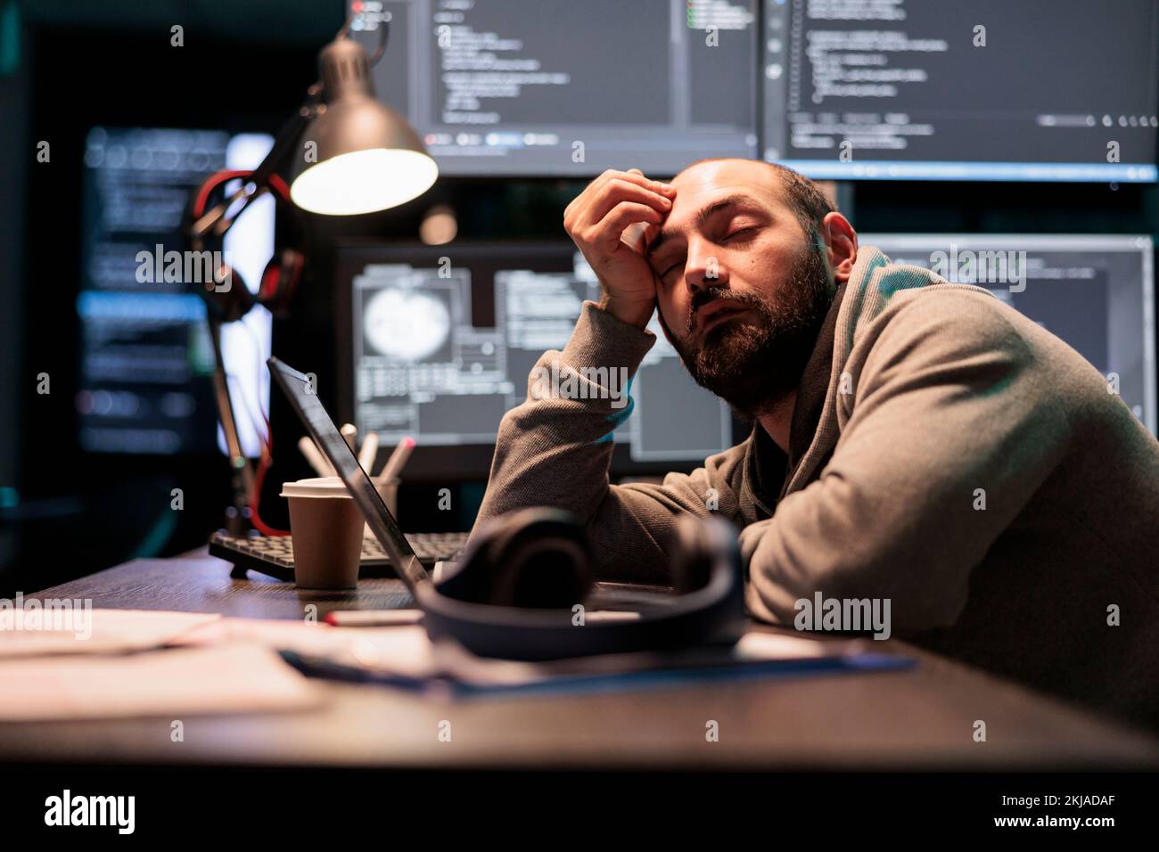 Exhausted app developer feeling sleepy late at night, falling asleep in office writing code on terminal window. Tired overworked programmer working after hours with artificial intelligence. Stock Photo