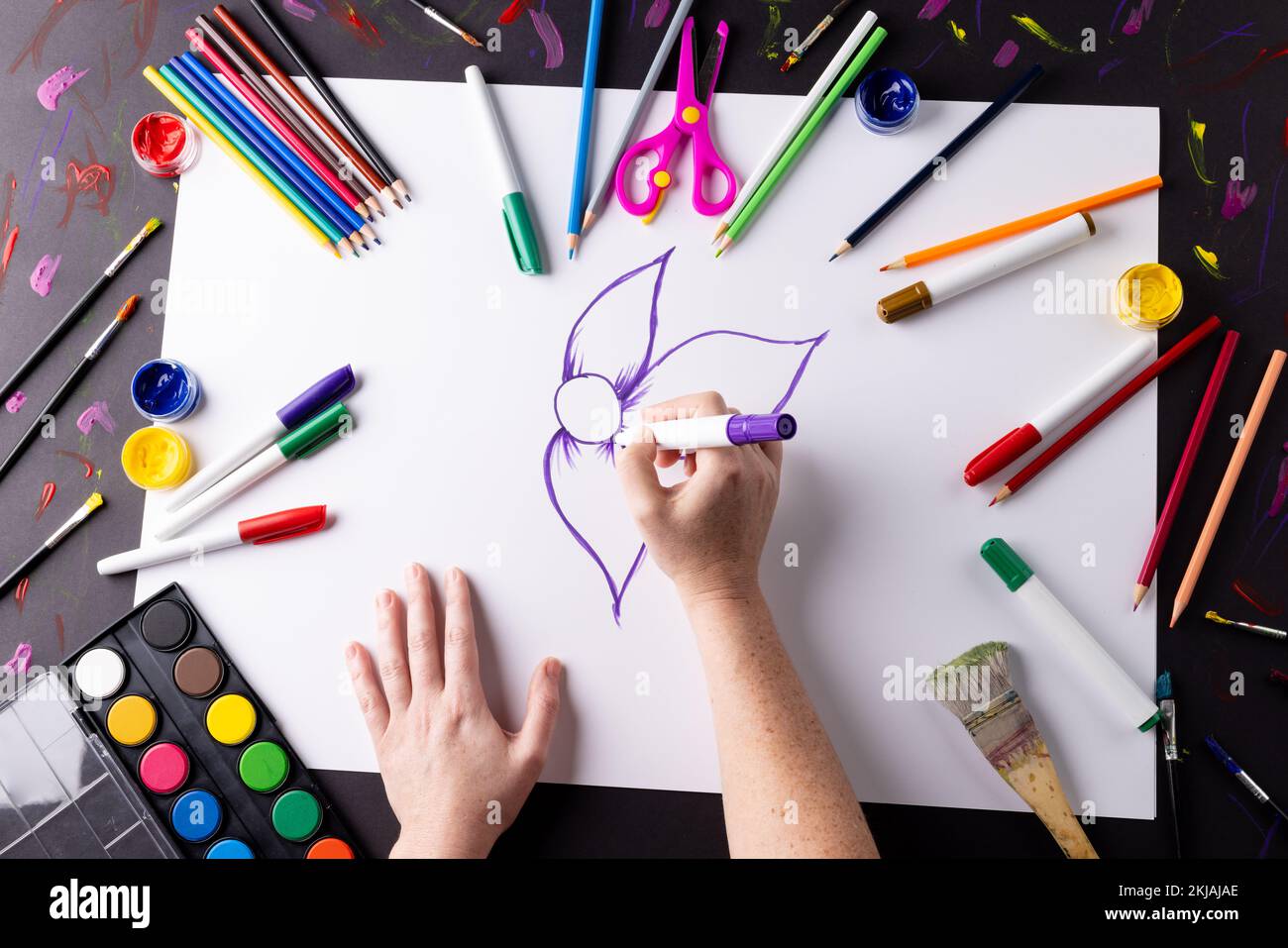 Overhead of hands drawing purple flower on paper, with art materials on table top Stock Photo