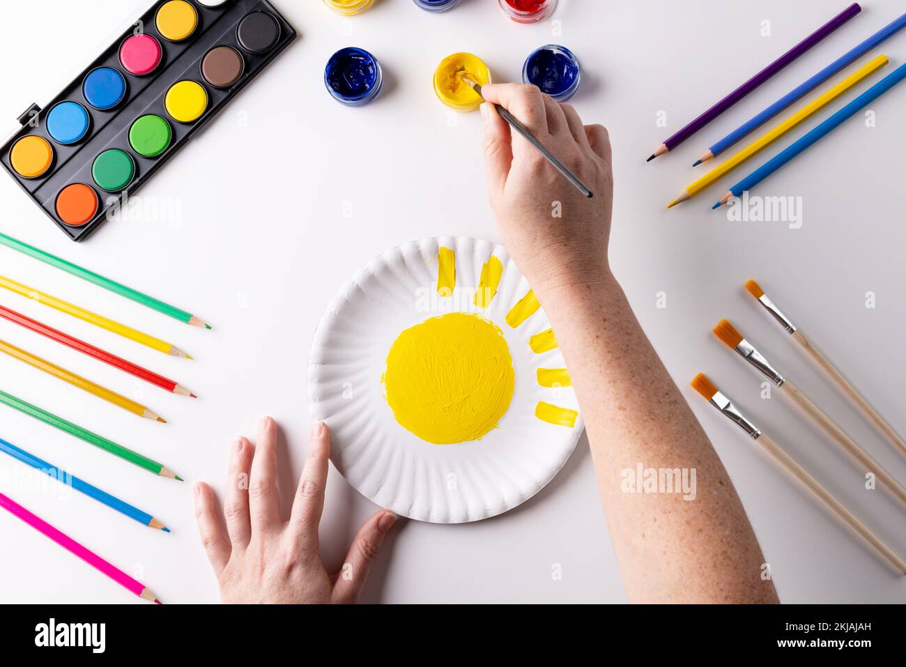 Overhead of hands painting with yellow paint on paper plate, with art materials on table top Stock Photo