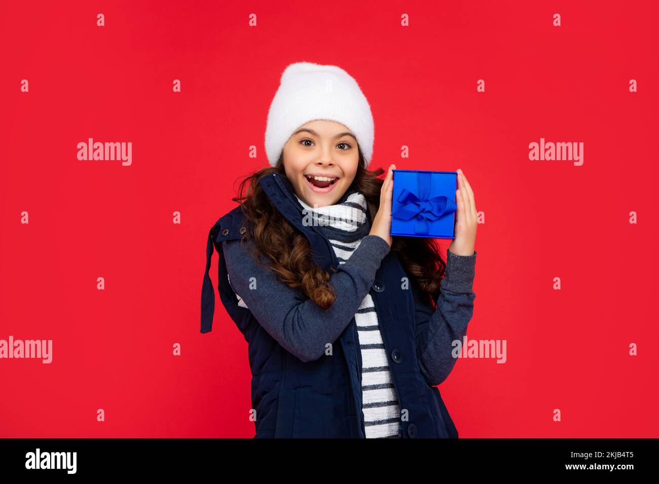 surprised child in puffer jacket and hat hold box. kid with present. teen girl on red background Stock Photo