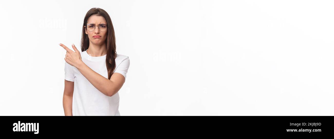 Waist-up portrait of moody, disappointed and complaining cute brunette woman in glasses, frowning and grimacing, sulking defensive, pointing finger Stock Photo