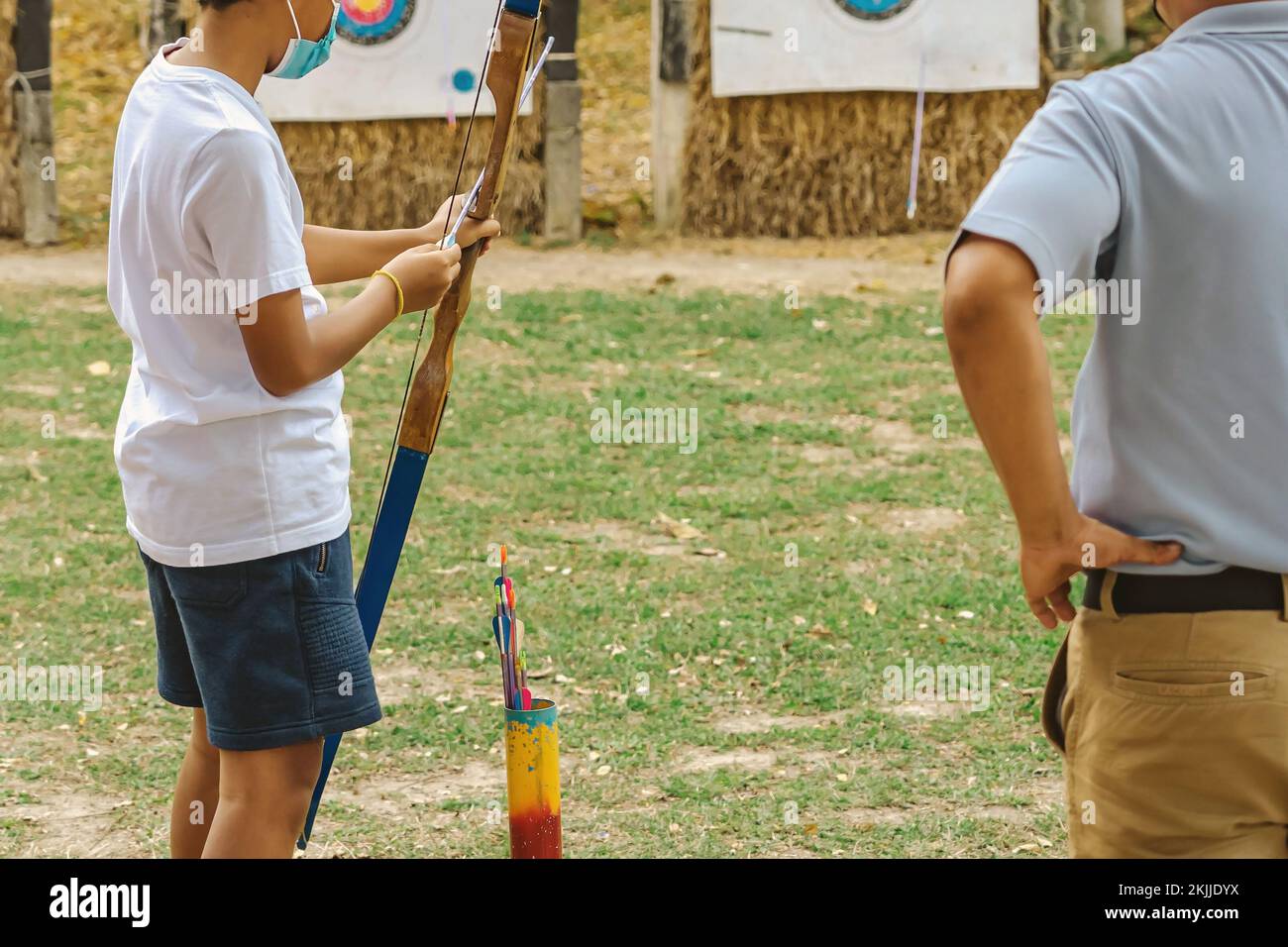 Back view of Asian boy wear face mask aims archery bow and arrow to colorful target in shooting range during training and competition. Exercise and co Stock Photo