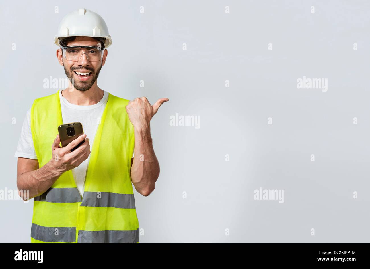 Smiling engineer holding phone and pointing to the side isolated. Young construction engineer holding phone and pointing at blank space. Civil enginee Stock Photo