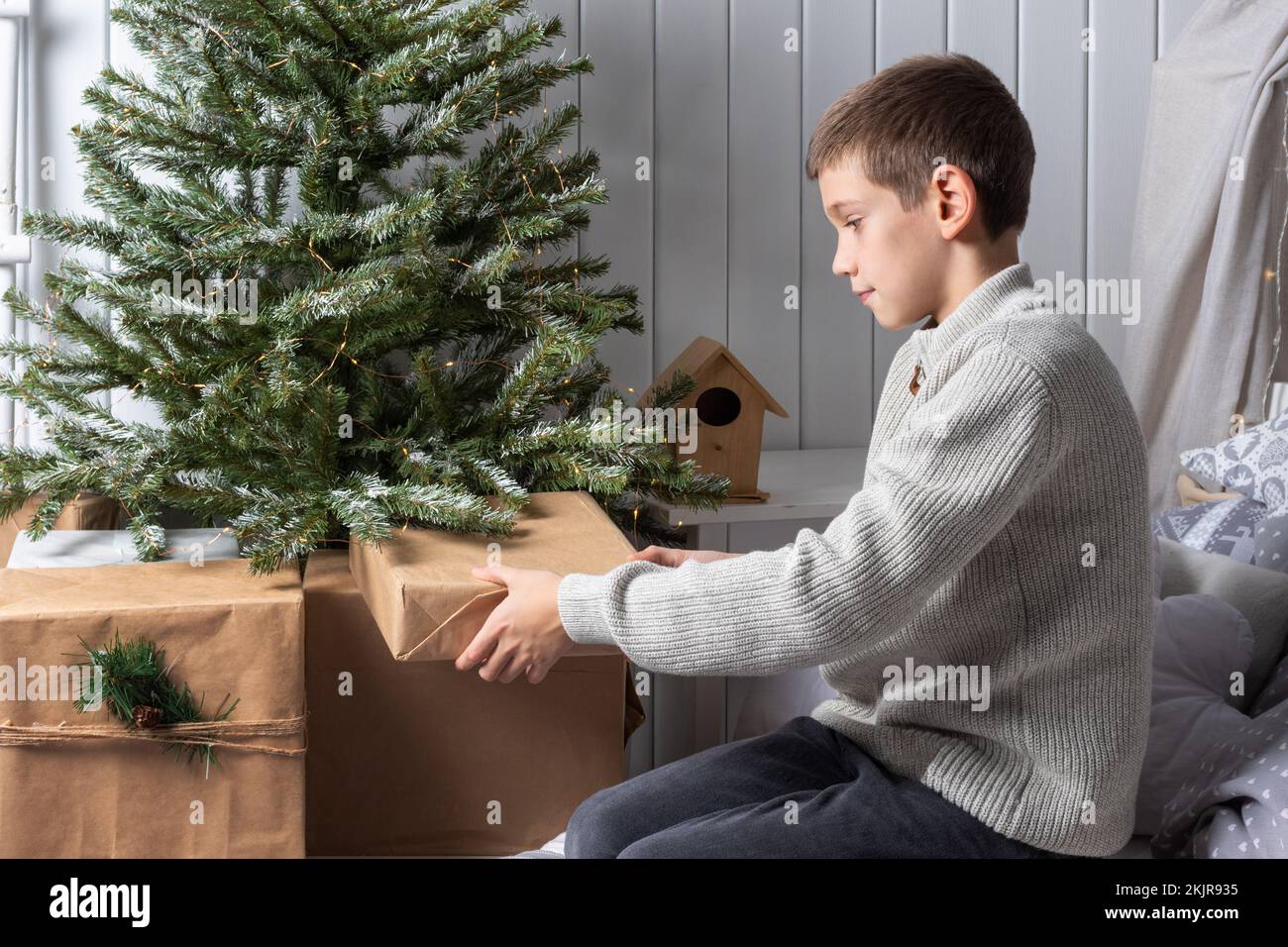 Cute teenage boy in a sweater sitting on the bed and putting presents under the Christmas tree. New Year's morning. Christmas Family Gifts Stock Photo