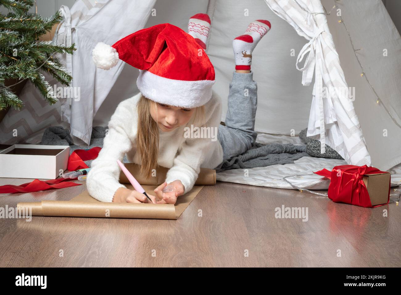 Christmas miracle wish list. Christmas helper child writing letter to Santa Claus in red hat. smiling girl making wish list or letter to santa at home Stock Photo