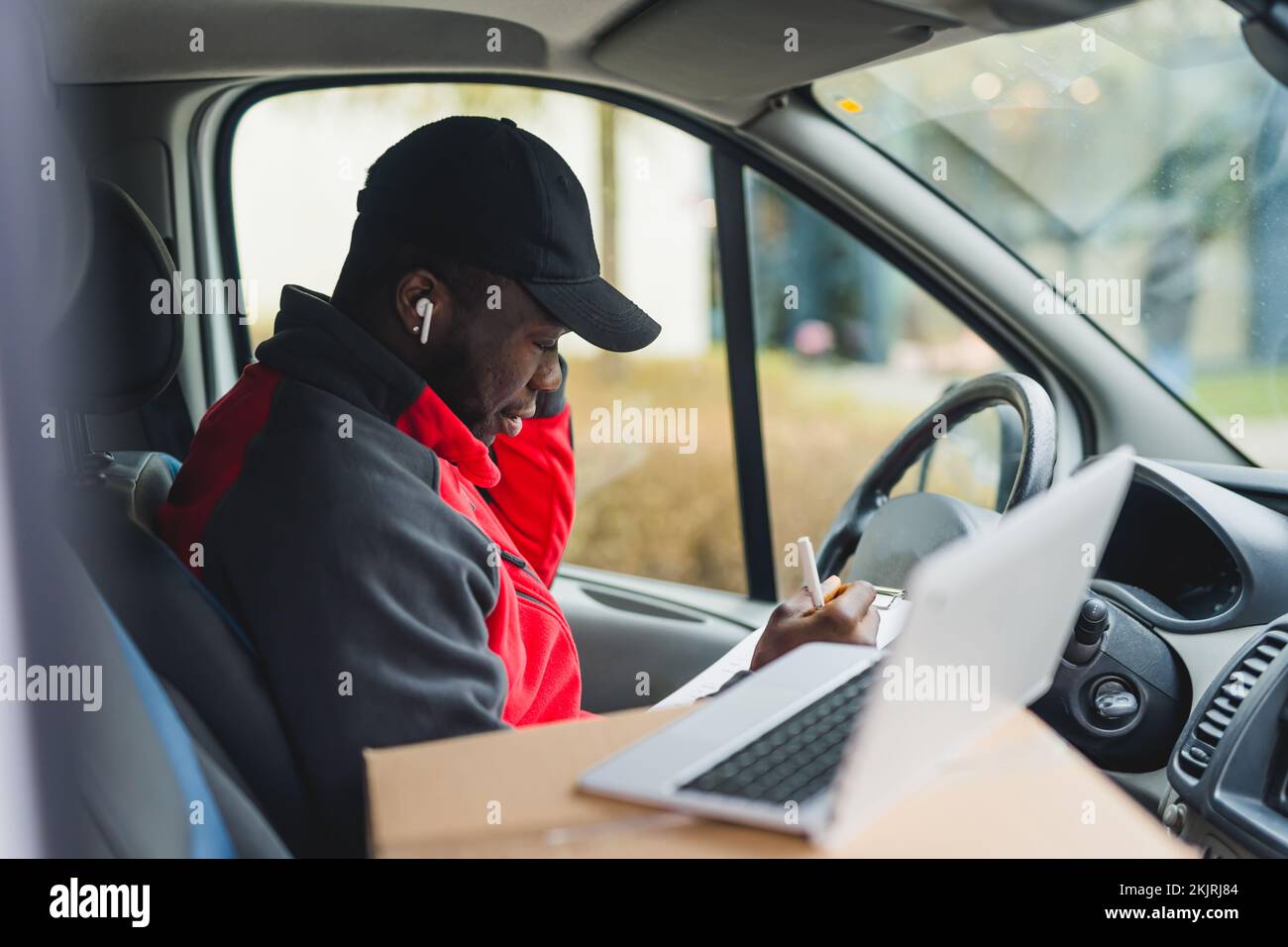 Black young adult delivery man sitting in driver seat of van filling out documents and working on laptop computer. Horizontal shot. High quality photo Stock Photo