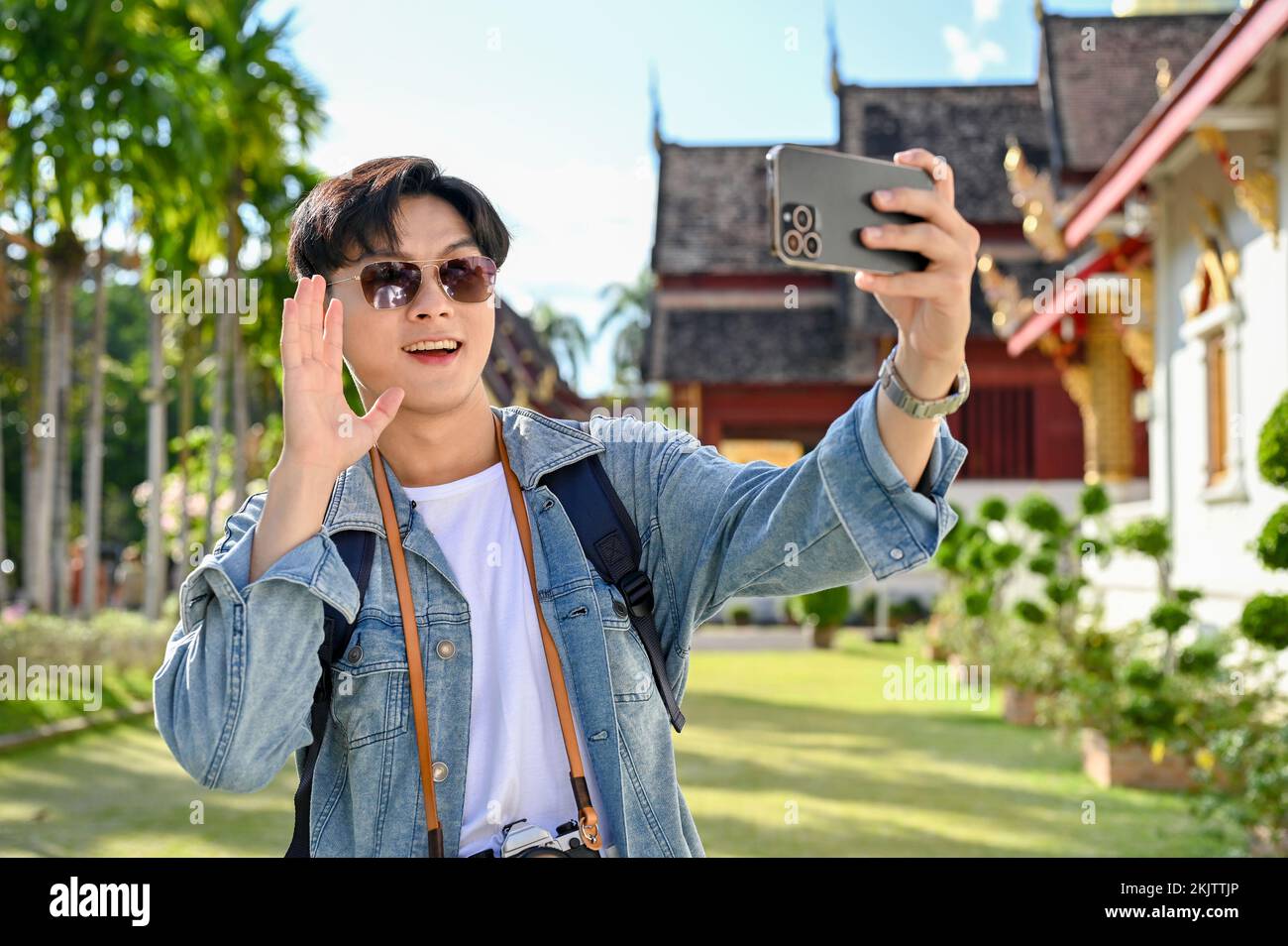 Handsome and cheerful young Asian male traveler wearing sunglasses, taking selfie or making a video vlog with his smartphone in the beautiful Thai's t Stock Photo