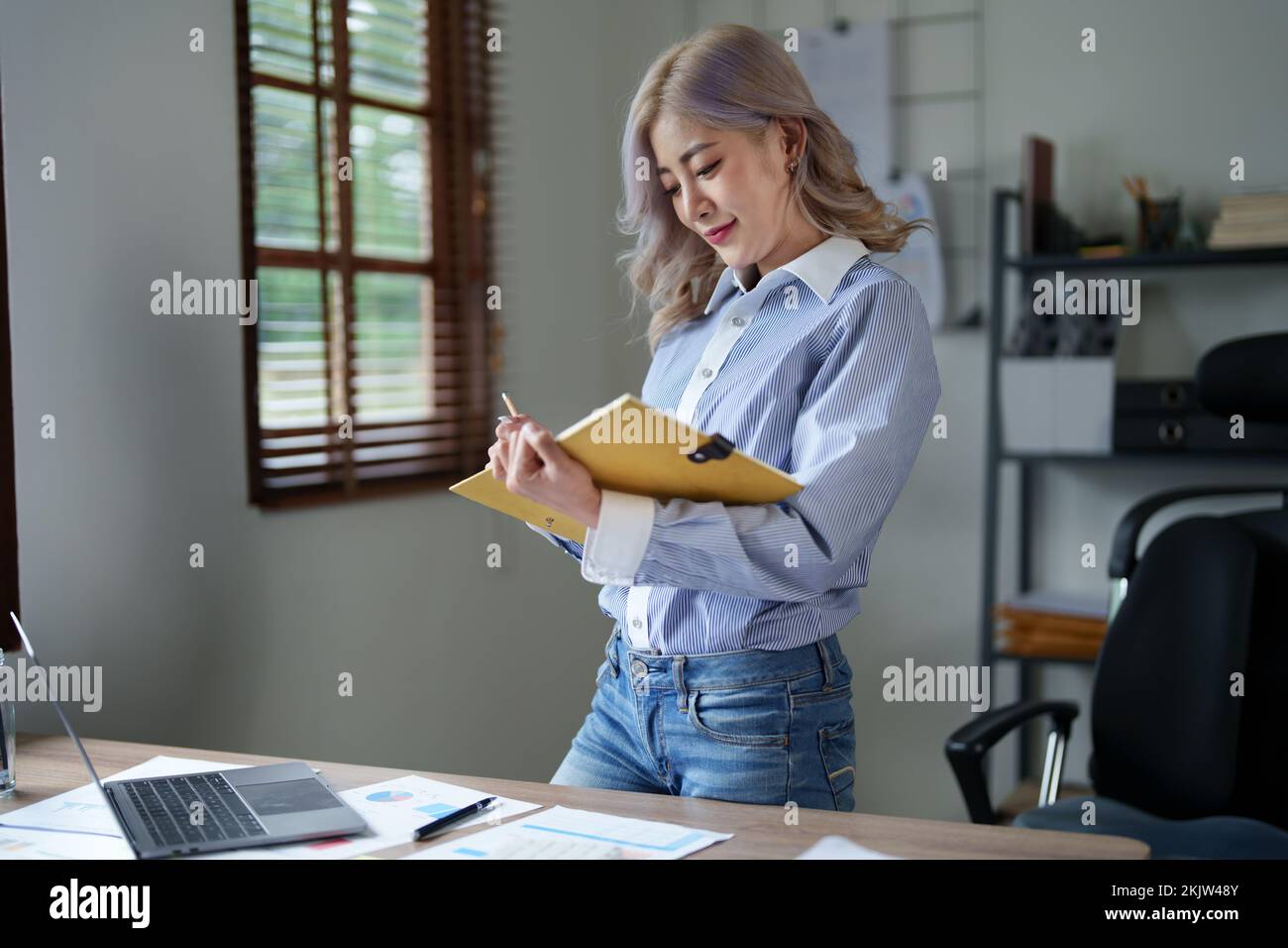 Portrait of a young Asian woman showing a smiling face as she using notebook, computer and financial documents on her desk in the early morning hours Stock Photo