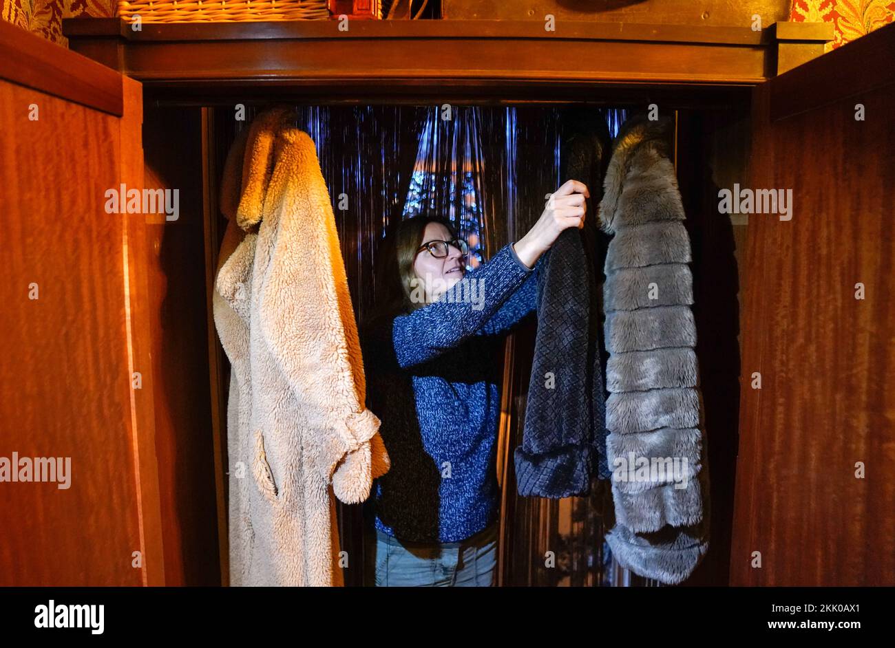A staff member at the National Trust's Mottisfont near Romsey in Hampshire, adjust coats hanging up in a wardrobe acting as a passage through to other rooms inside the National Trust's Mottisfont which have been transformed into scenes from The Lion, the Witch and the Wardrobe during a Christmas exhibition preview. Picture date: Friday November 25, 2022. Stock Photo