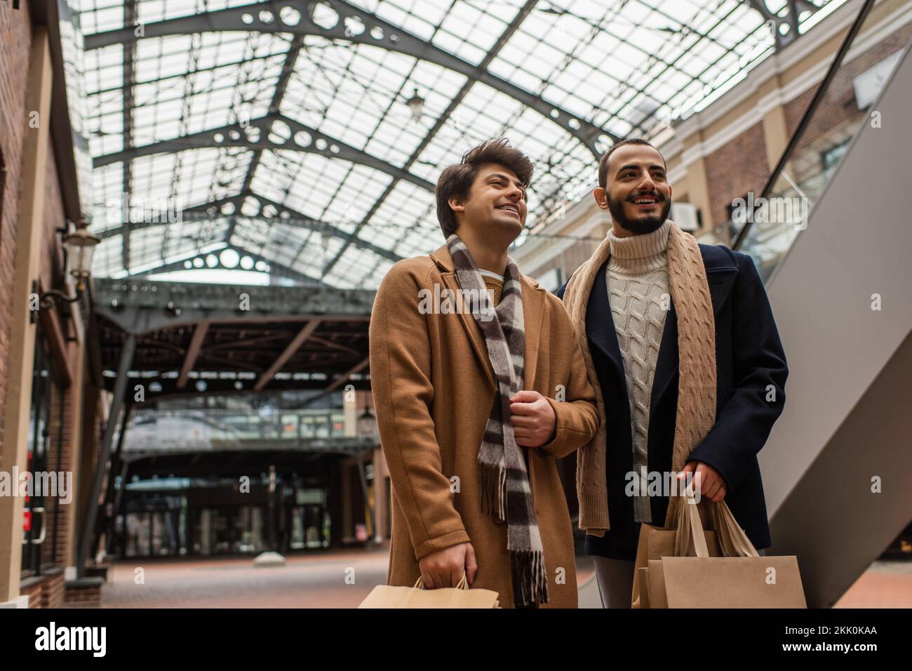 cheerful gay boyfriends in trendy clothes standing with shopping bags under transparent roof on street,stock image Stock Photo