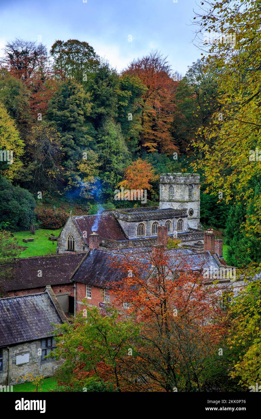 Spectacular autumn colour on the woodland trees, a smoking chimney and the parish church at Stourhead Gardens, Wiltshire, England, UK Stock Photo