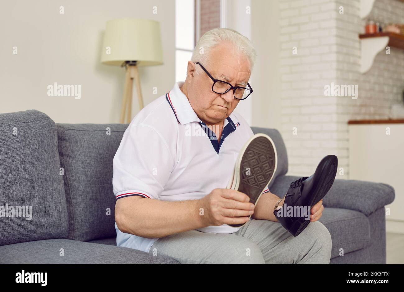 Demented retired senior man sitting on couch and choosing which type of shoes to wear Stock Photo