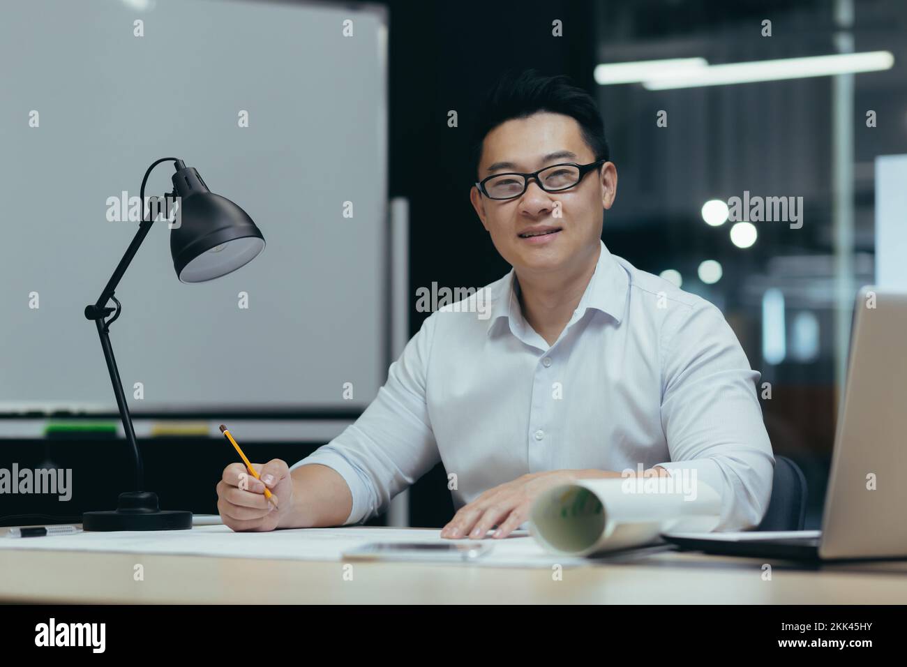 Portrait of a young handsome Asian man, architect, designer, freelancer. Sitting in the office at the table in glasses with a laptop. Makes a drawing, makes a plan. He looks at the camera, smiles. Stock Photo