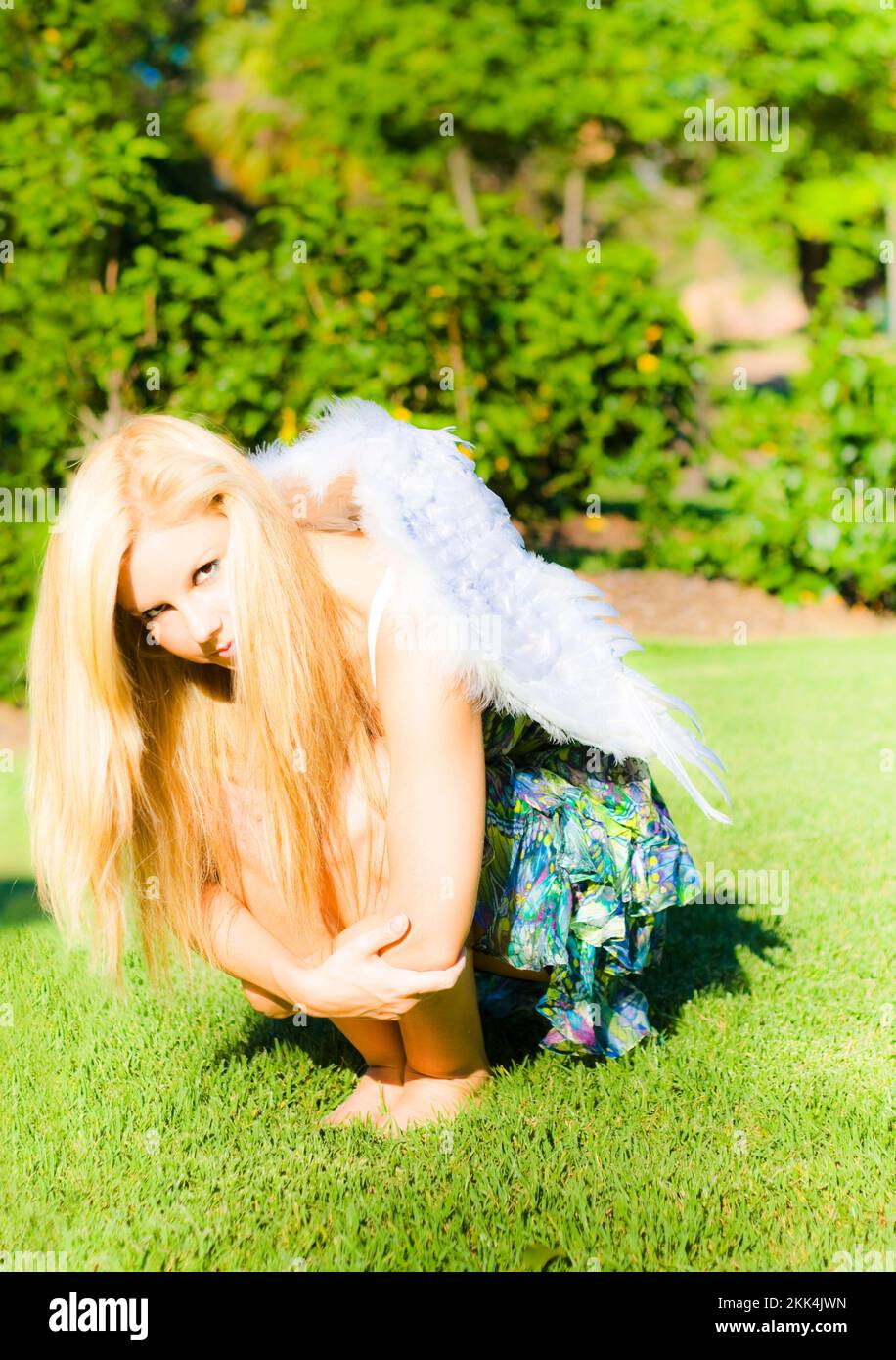 Woman with long blonde hair squatting arms around her knees, with feather wings on her back in a broken wings concept Stock Photo