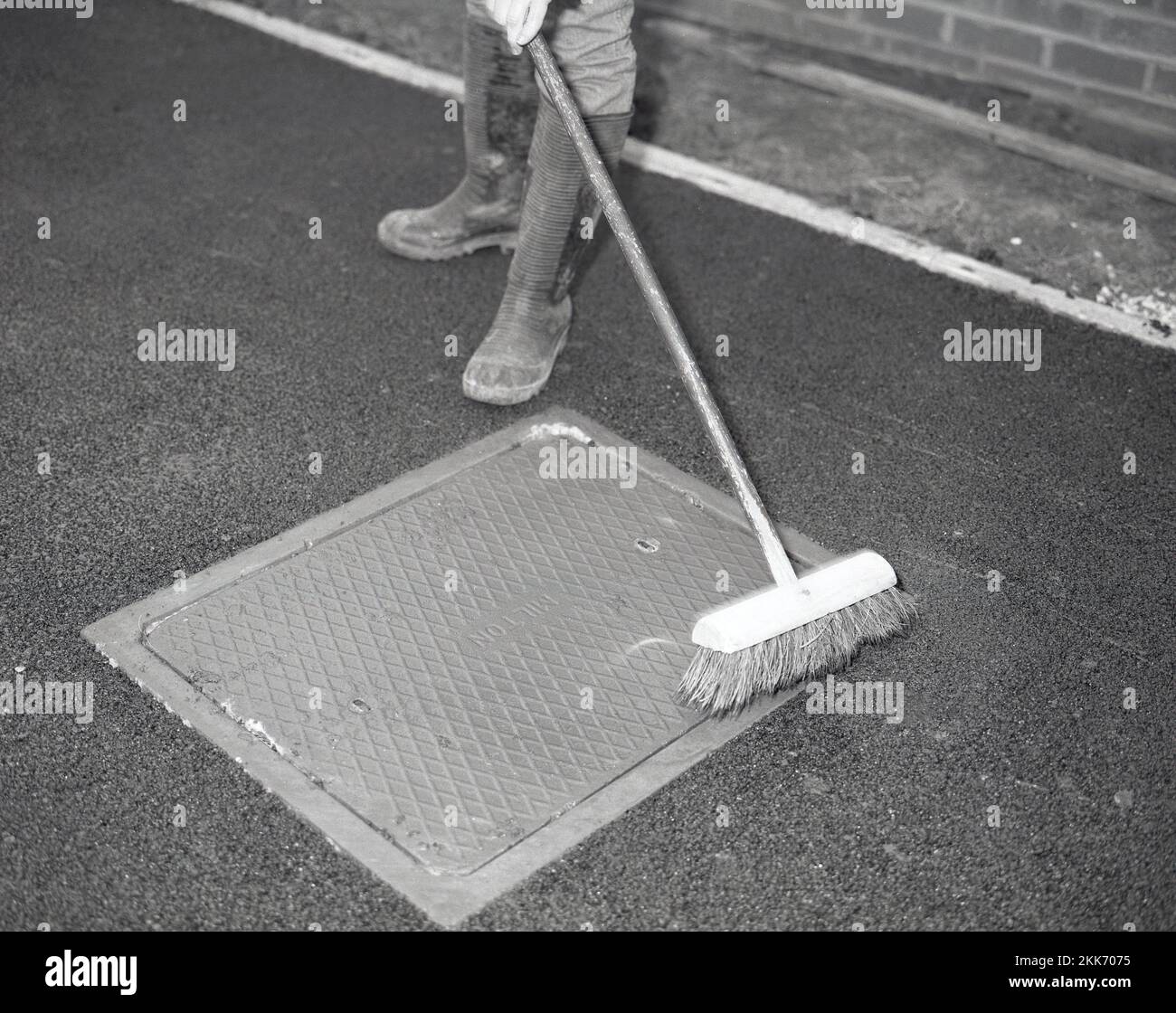 1980s, re-surfacing a driveway, a worker using a broom to brush any black macadam residue left on a manhole cover after asphalting a household driveway, England, UK. Stock Photo