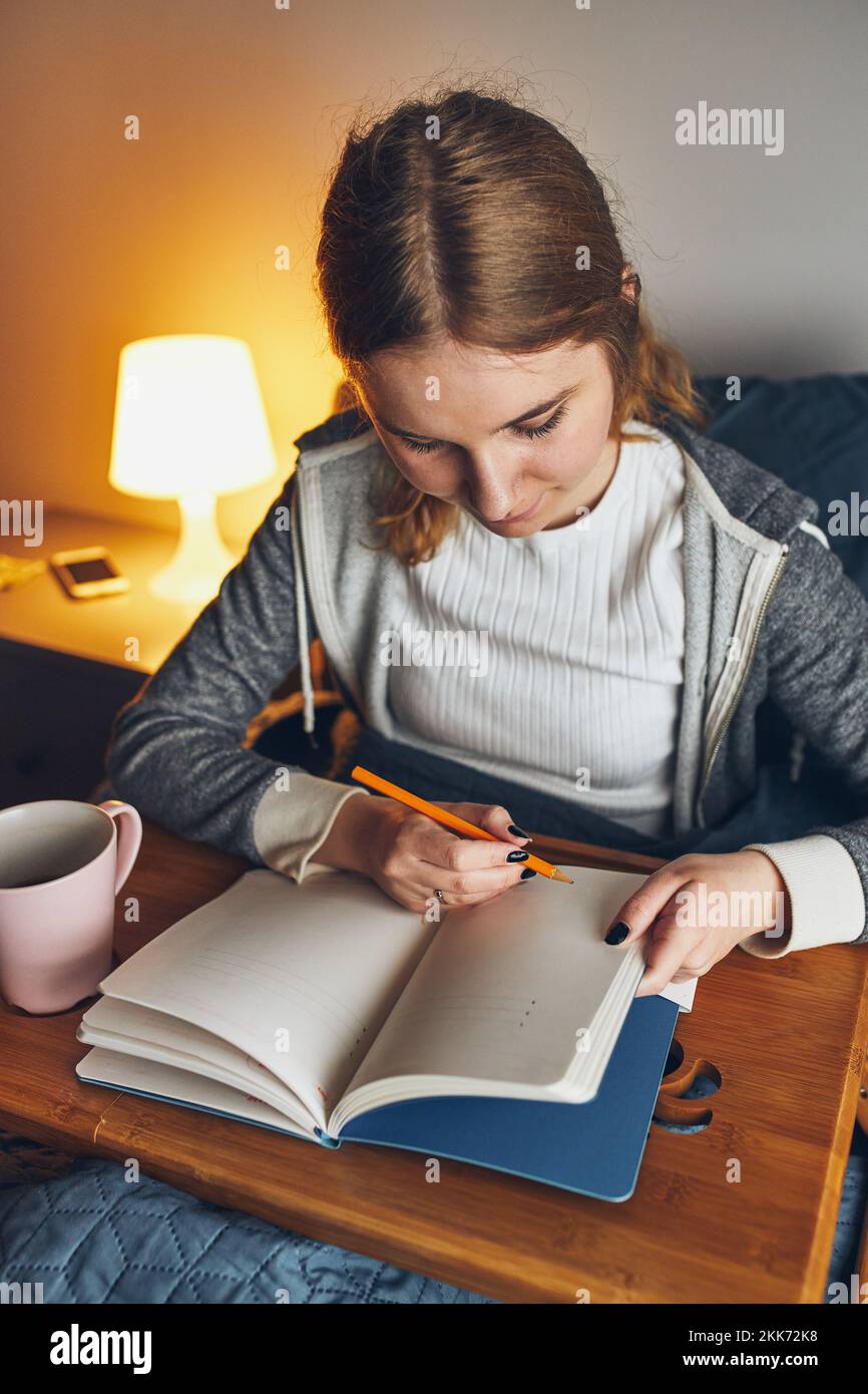 Student learning at home. Young woman making notes, reading and learning from notepad. Girl writing journal sitting in bed at home during quarantine Stock Photo