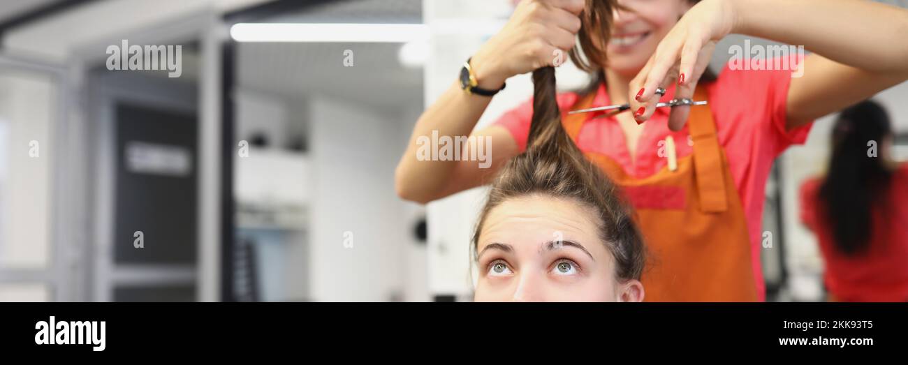 Hairdresser going to cut lock of long hair with scissors tool, female client scared in masters chair Stock Photo