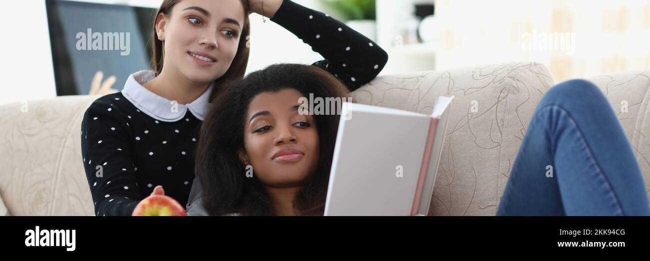Best friends reading book chilling on sofa, apple for snack, relaxing atmosphere Stock Photo
