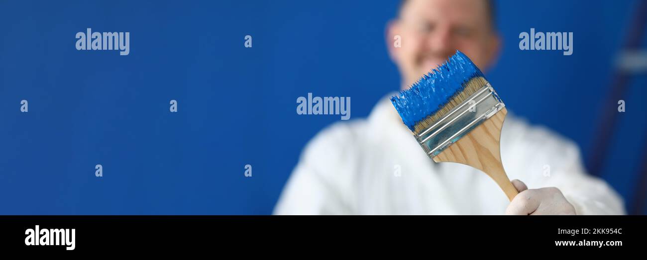 Painter worker hold brush covered in blue colour paint, handyman on construction site Stock Photo