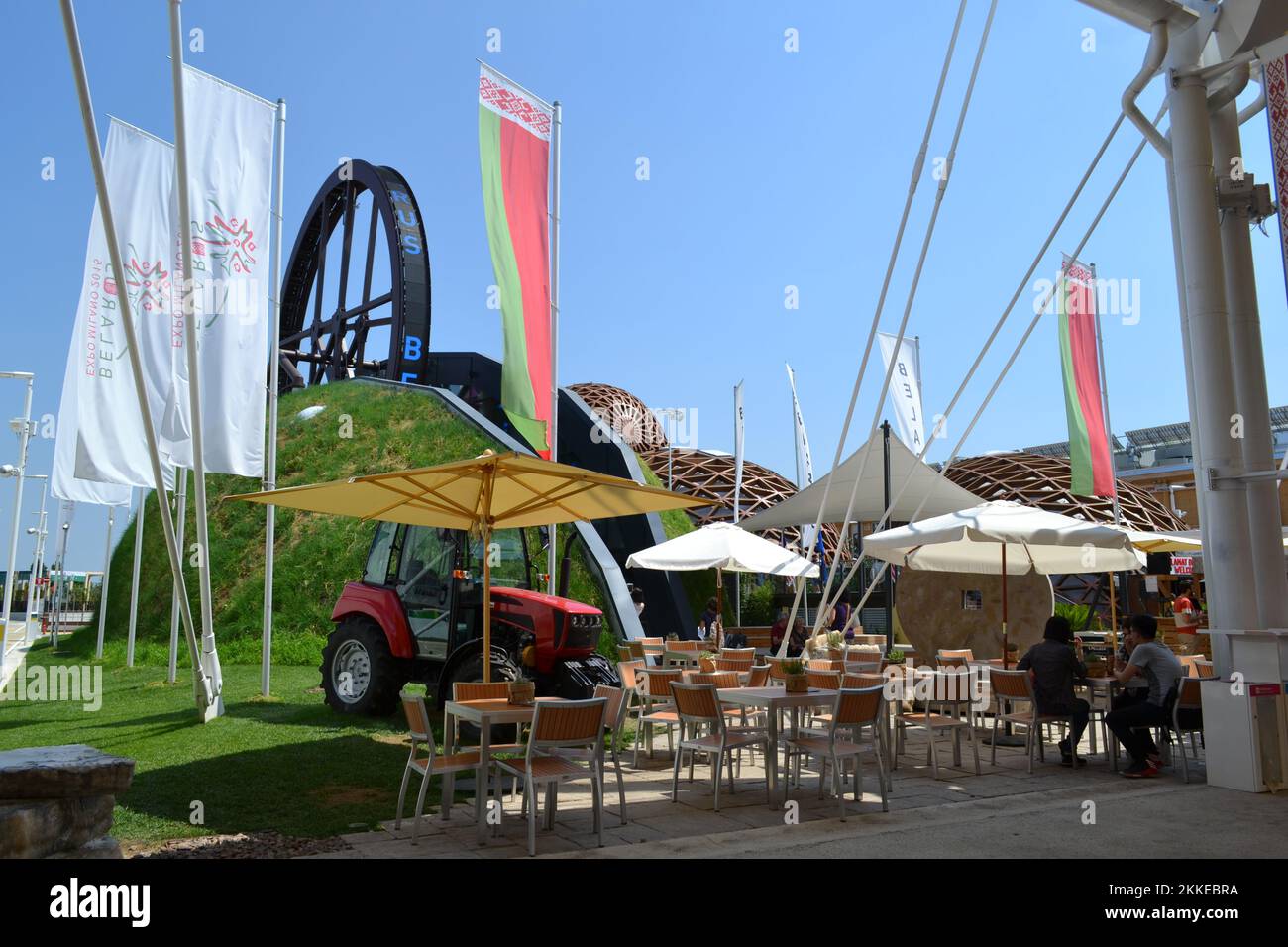 Panoramic view of the Belarus pavilion outdoors at Expo Milan 2015. Bar area with umbrellas. Giant wheel - 'Wheel of life' - symbol of l food chain. Stock Photo