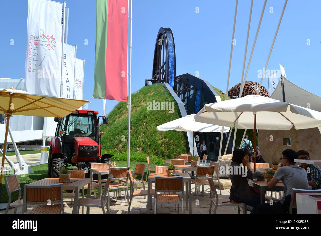 Panoramic view of the Belarus pavilion outdoors at Expo Milan 2015. Bar area with umbrellas. Giant wheel - 'Wheel of life' - symbol of l food chain. Stock Photo