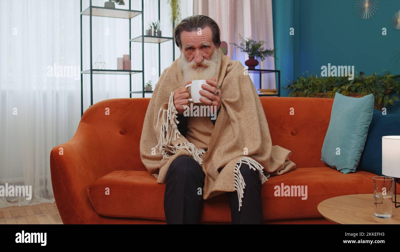 Sick senior grandfather man wrapped in plaid shivering from cold on sofa drinking hot tea in unheated apartment without heating due debt. Unhealthy elderly grandpa feeling discomfort try to warming up Stock Photo
