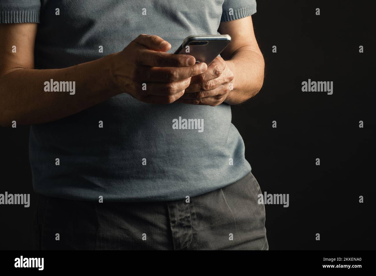 Cropped front view of a man wearing jeans and short-sleeved shirt using smartphone, indoor environment with black backdrop. Male hands with cell phone Stock Photo