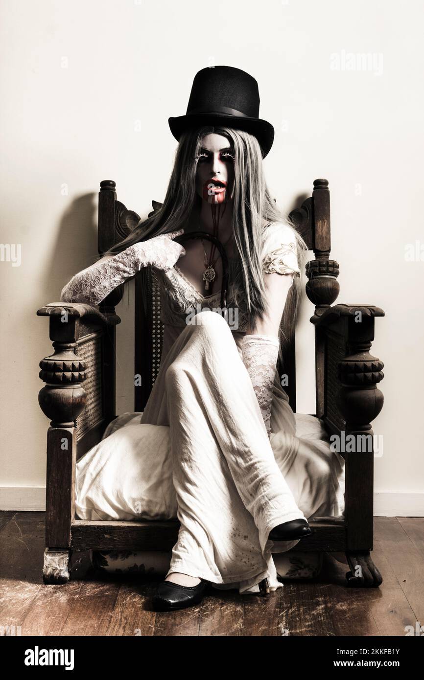 Scary horror image of a spooky elegant fashionable vampire woman in top hat and white dress sitting in a vintage armchair with bloody mouth Stock Photo