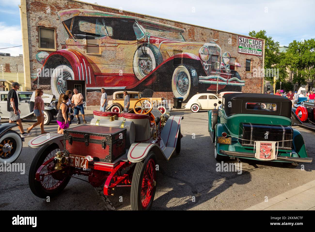 Classic cars on display beneath a mural depicting an antique Duesenberg