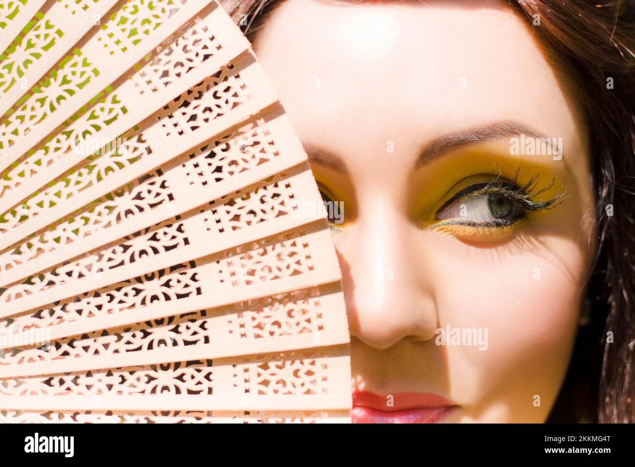 A Beautiful Mischievous Woman Wearing Yellow Eyeliner Makeup Hides With A Sly And Sneaky Smirk Behind A Wooden Hand Fan In A Playful Fun In The Sun Im Stock Photo