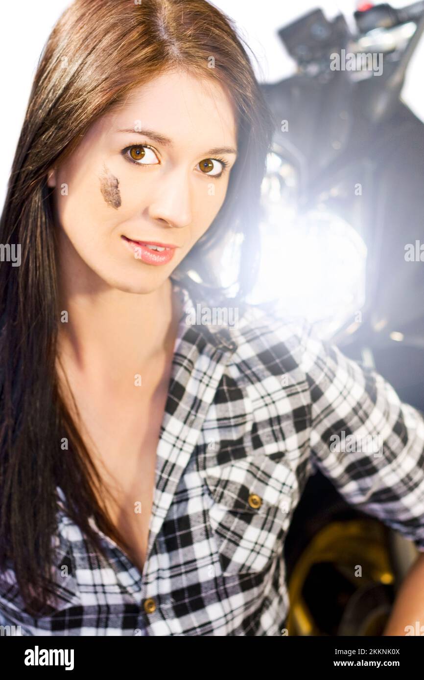 Female Motorcyclist Concept. Attractive young brunette woman standing facing camera with the bright headlight of a motorcycle shining over her left sh Stock Photo