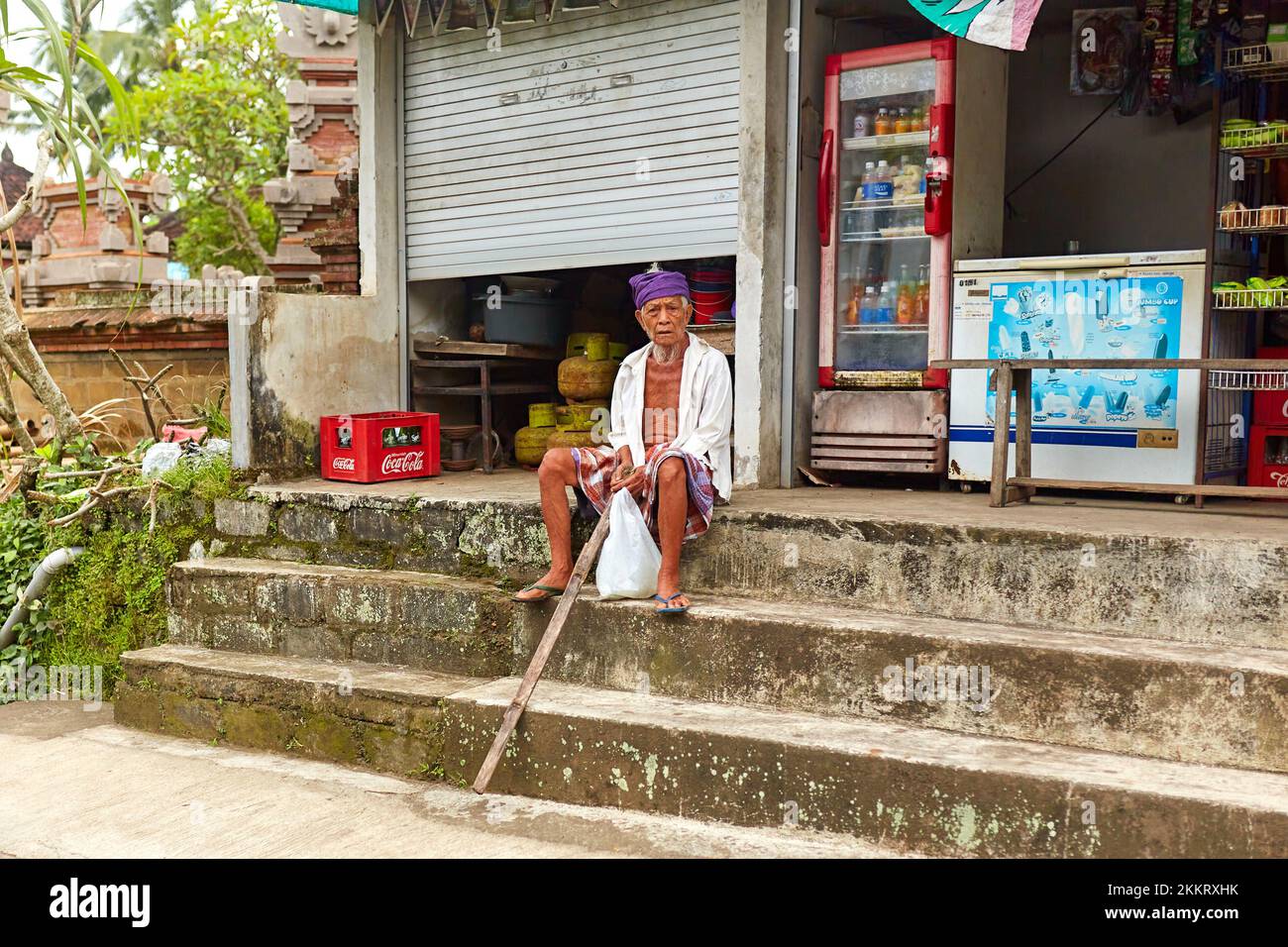 An elderly man in a sterno sits on the steps outside a grocery store. Bali, Indonesia - 03.22.2018 Stock Photo