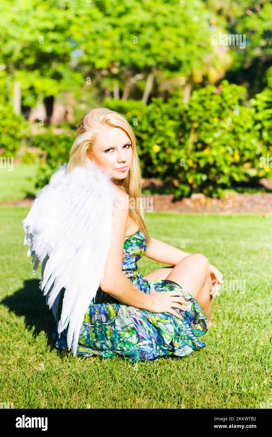 Beautiful young celestial angel in white wings seated on the grass looking back over her shoulder at the camera with a gentle expression Stock Photo
