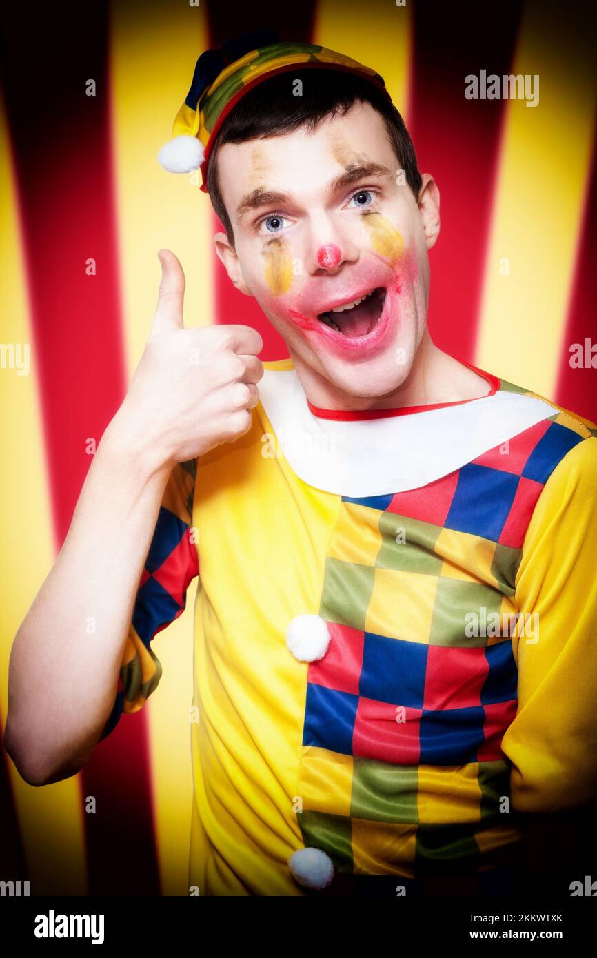 Playful Smiling Circus Clown Standing Inside Bigtop Tent Giving Thumbs Up For Good Entertainment While Gesturing A Trapeze Act Above Stock Photo