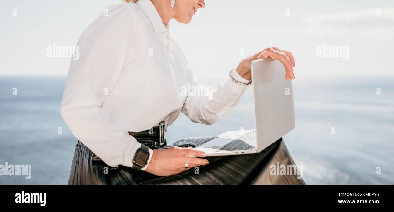 Digital nomad, Business woman working on laptop by the sea. Pretty lady typing on computer by the sea at sunset, makes a business transaction online Stock Photo