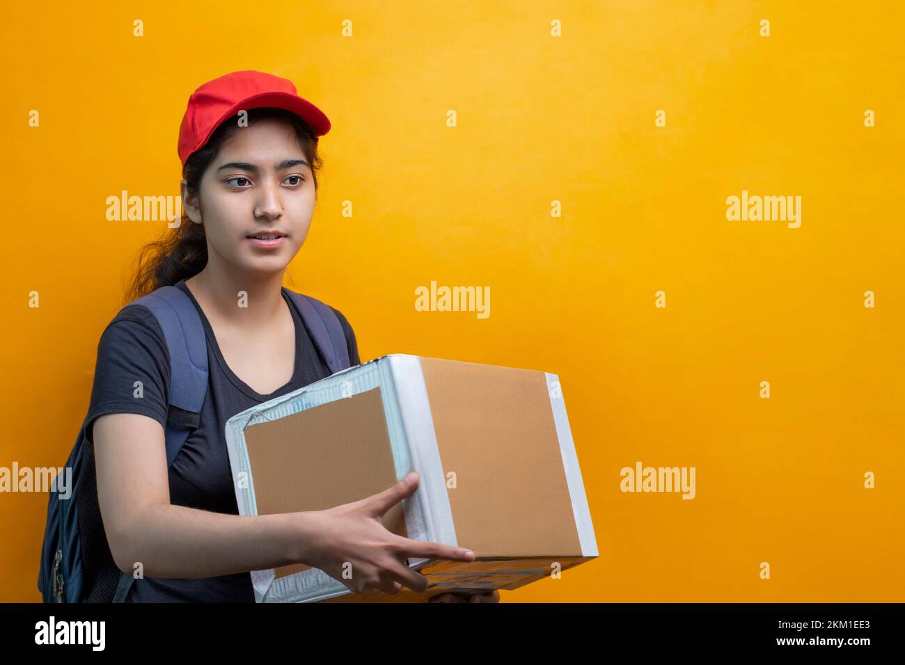Young Delivery Woman Holding Cardboard Box Against Yellow Background Stock Photo