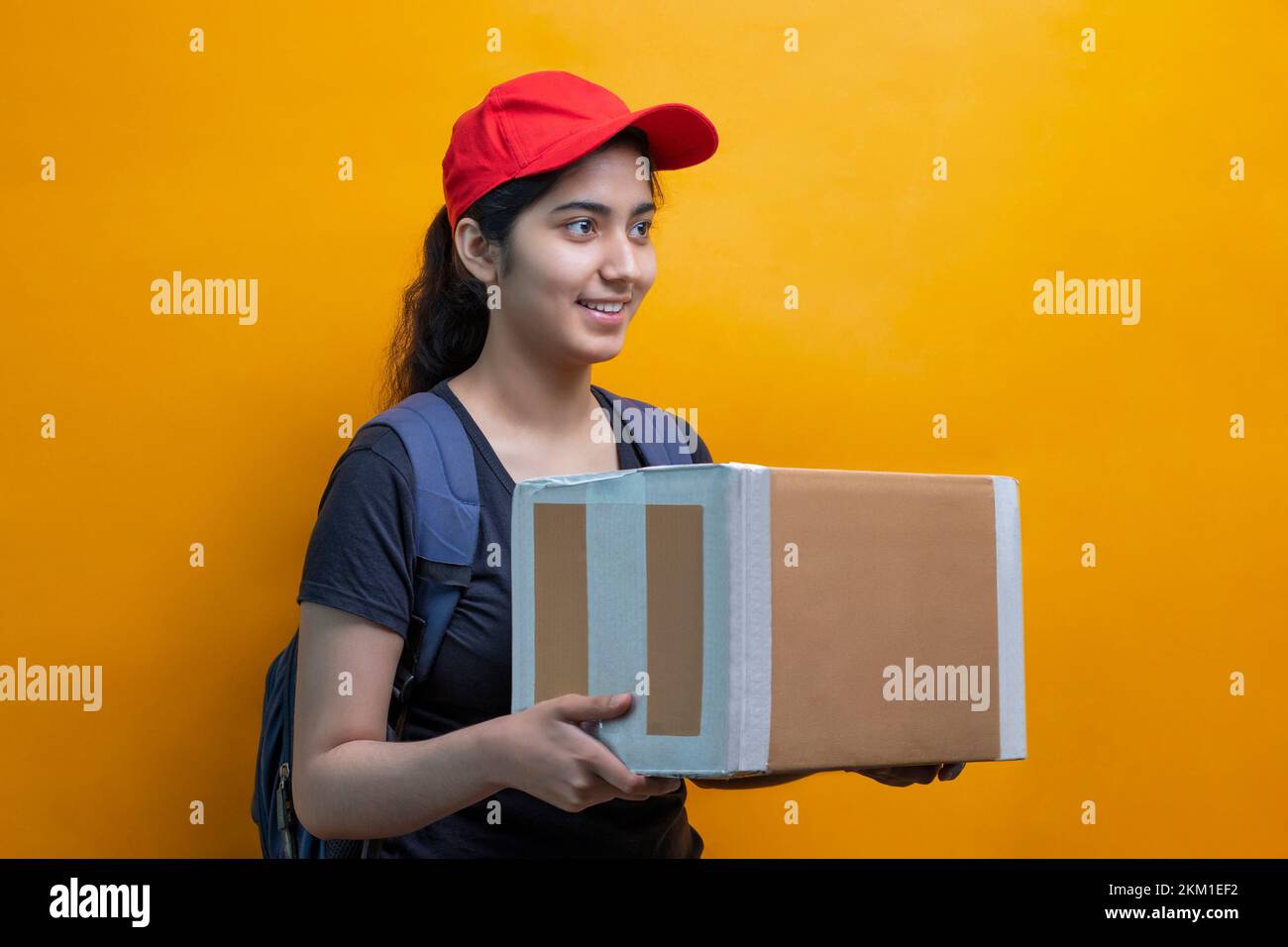 Young Delivery Woman Holding Cardboard Box Against Yellow Background Stock Photo