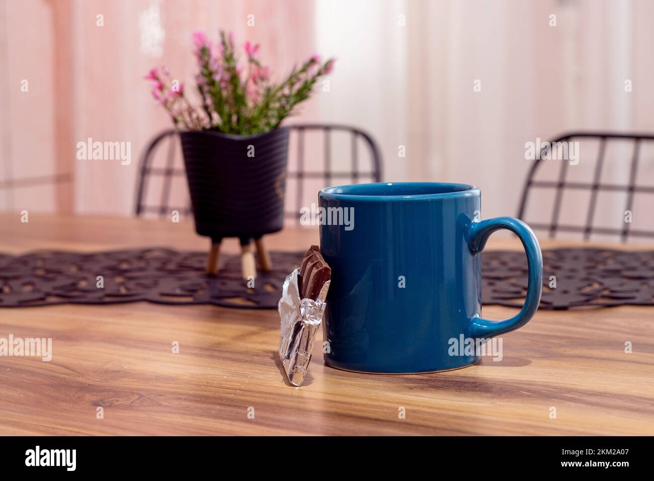 Coffee mug and chocolate on wooden table against curtain and chair background. Side view, copy space, close-up. Kitchen dining table, break concept Stock Photo