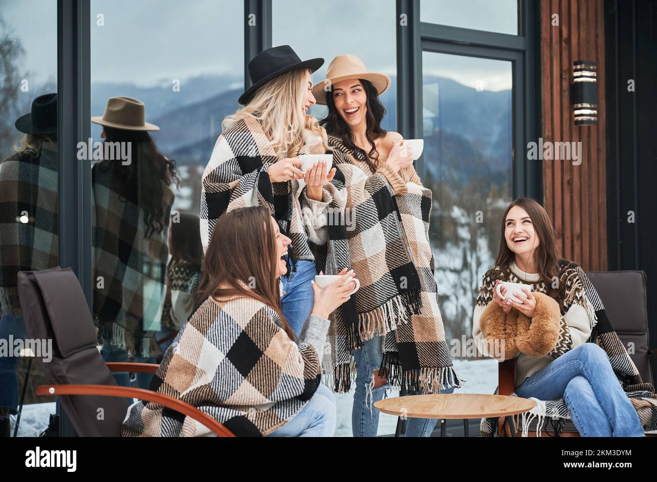 Young women enjoying winter weekends on terrace of contemporary barn house. Four girls having fun, sitting on chairs and drinking hot tea. Stock Photo