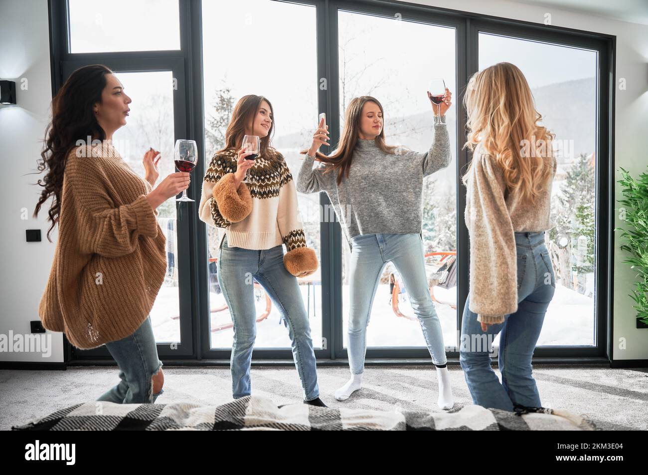 Young women enjoying winter weekends inside contemporary barn house. Four girls having fun and drinking red wine. Stock Photo