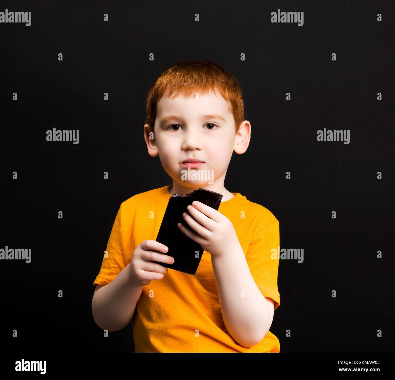 portrait the face of a small red-haired boy bites and bites a bar of sugar chocolate, close-up portrait of a child with sweets Stock Photo