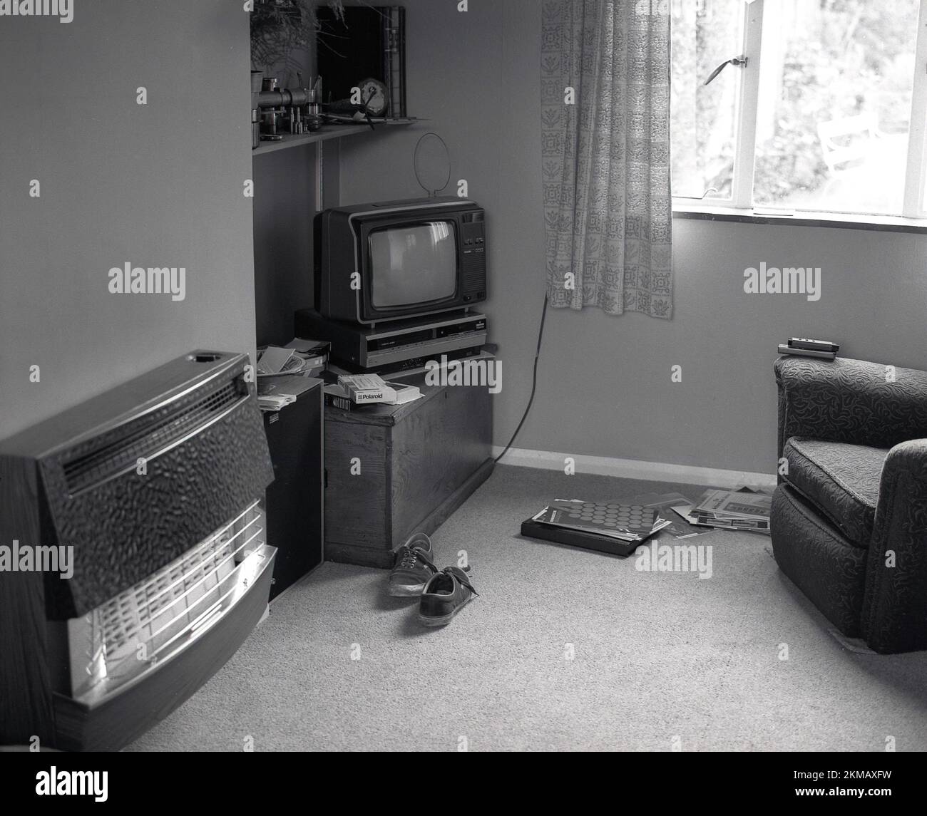 1980s, historical, inside a small living room, picture shows a wall-mounted brass fronted gas fire, wooden chest with a small television of the era on it with wire ariel and underneath it, a video player and videos. Opposite a sofa chair with televison controls on arm, England, UK. Stock Photo