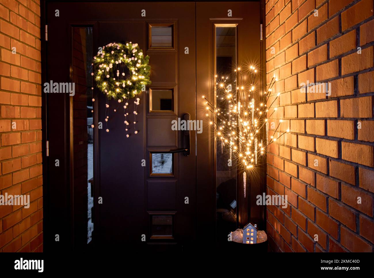 Home door entrance outdoors with various Christmas decorations Christmas wreath with pom poms and flower pot with artificial tree with lights. Stock Photo