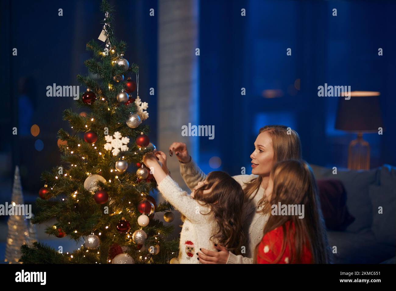 Pretty caucasian woman and little girls decorating christmas tree with colorful toys. Young mother with two daughters spending evening time at home wi Stock Photo