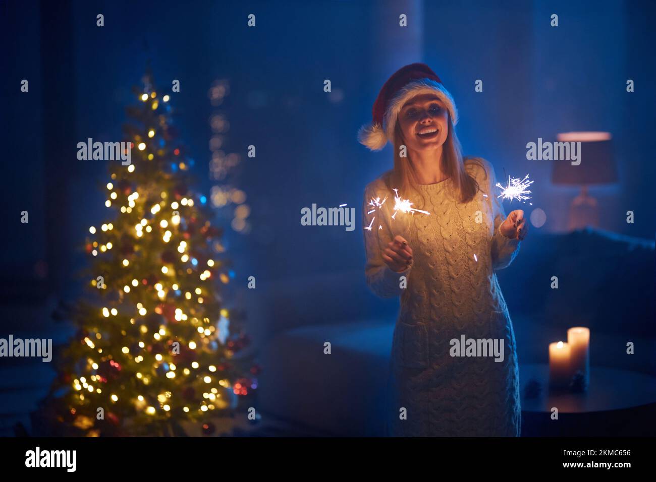 Smiling young lady on knitted dress and santa hat standing near shiny christmas tree with sparklers in hands. Charming caucasian blonde celebrating wi Stock Photo
