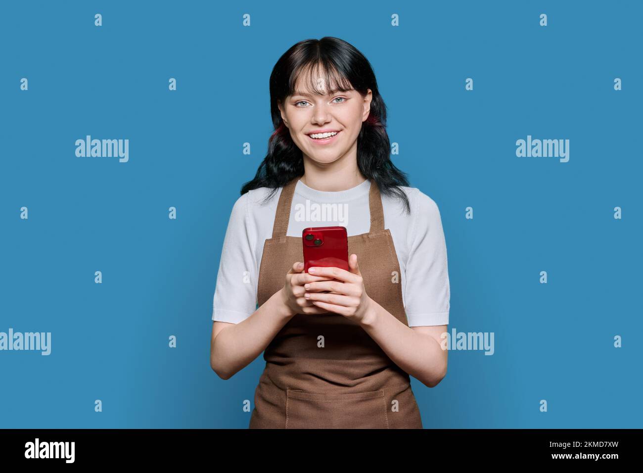 Smiling young female staff in apron using smartphone, on blue background Stock Photo