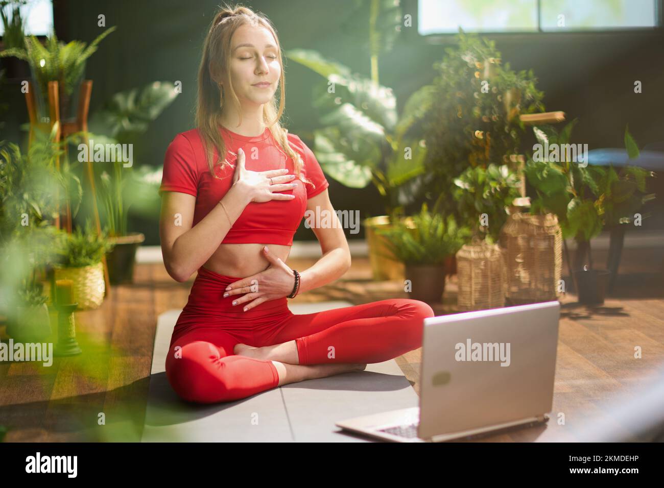 modern woman in red fitness clothes with yoga mat and laptop doing yoga in the modern green living room. Stock Photo