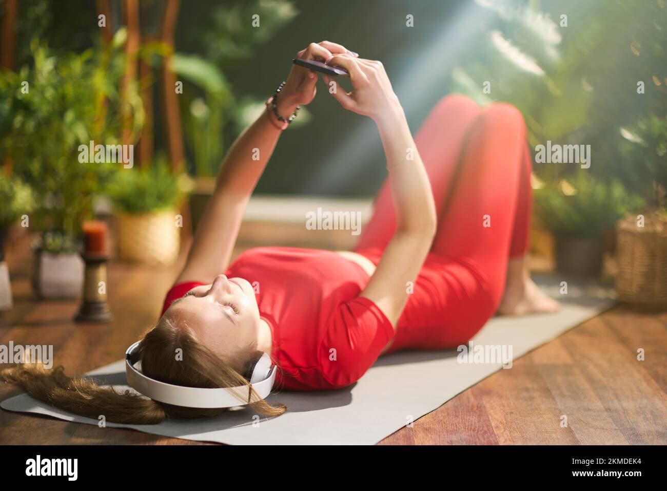 young female in red fitness clothes with yoga mat using smartphone and listening to the music with headphones at modern green home. Stock Photo